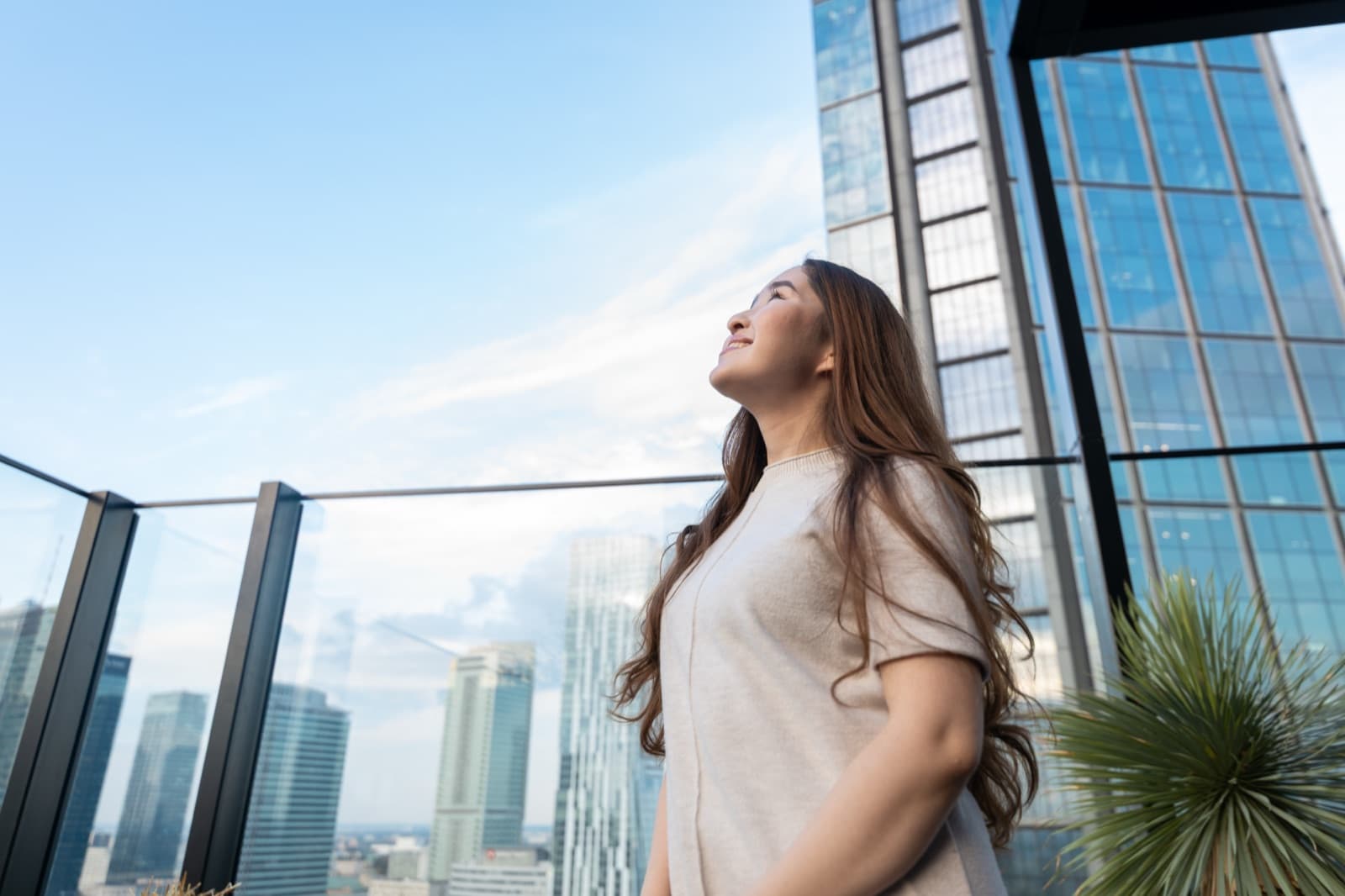 A woman on a city rooftop terrace gazing up at the sky between tall glass buildings